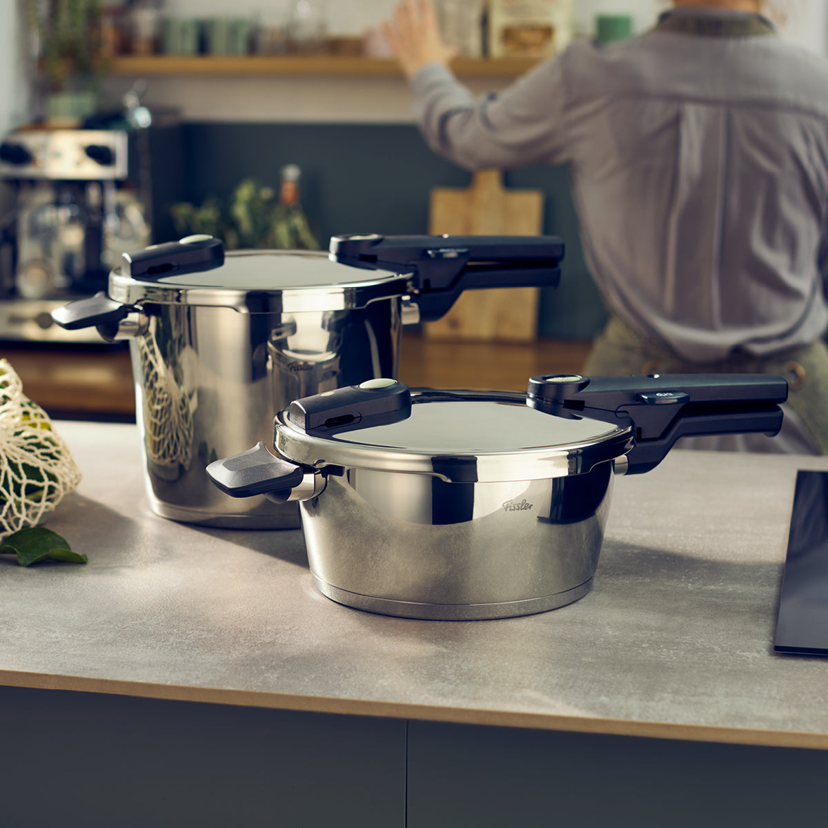 Zwei Fissler Schnellkochtöpfe aus rostfreiem Stahl glänzen auf einem schlichten Küchentisch; eine Person bereitet die Speisen vor und präsentiert dabei deutsche Qualität und Design.