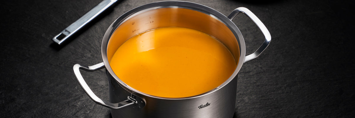 Ein in Deutschland präzisionsgefertigter Fissler-Edelstahltopf enthält eine orangefarbene Cremesuppe; dahinter befindet sich ein Schöpflöffelgriff aus Metall.