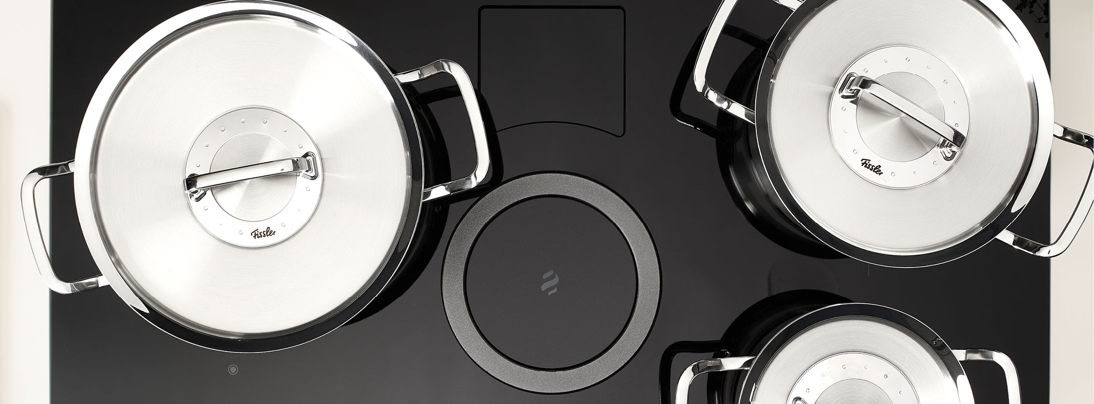 Drei Fissler-Edelstahltöpfe mit Deckeln auf einem eleganten schwarzen Glaskochfeld, das deutsche Präzision und zeitloses Design präsentiert.