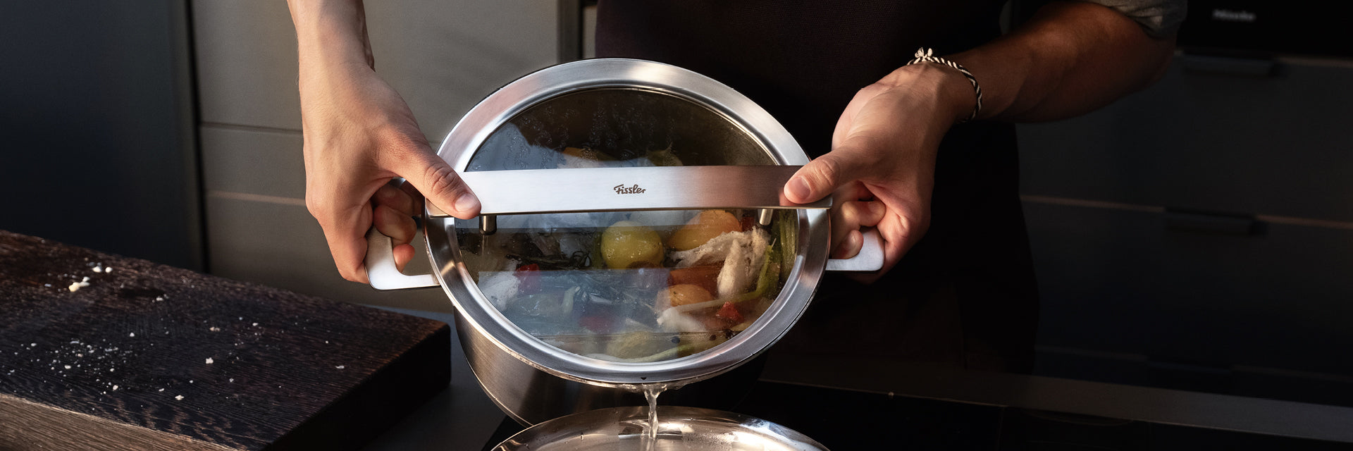 Eine Person gießt in einer eleganten, modernen Küche Wasser aus einem Fissler-Edelstahltopf mit Glasdeckel in eine Pfanne ab.