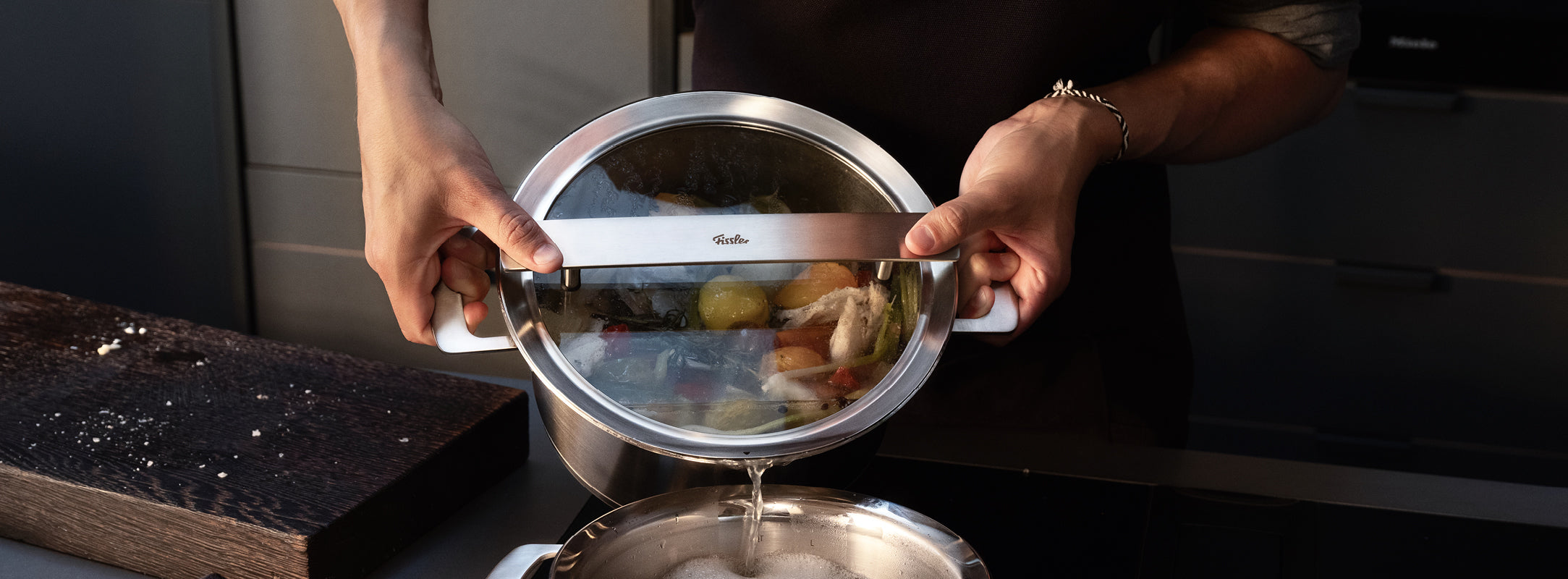 Eine Köchin gießt Gemüse mit einem Fissler Topf mit Präzisionssiebdeckel in einer eleganten, modernen Küche ab. Deutsche Qualität, zeitloses Design.