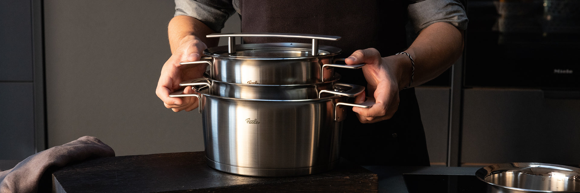 Eine Person in einer dunklen Schürze hält Fissler-Edelstahltöpfe mit Deckeln, die deutsche Qualität und zeitloses Design auf der Theke präsentieren.