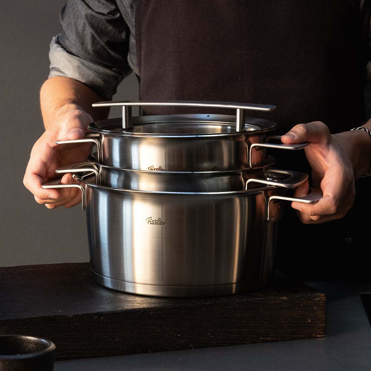 Eine Person in dunkler Schürze stapelt Fissler-Edelstahltöpfe mit Deckeln auf einer dunklen Holzoberfläche und zeigt zeitloses deutsches Design.