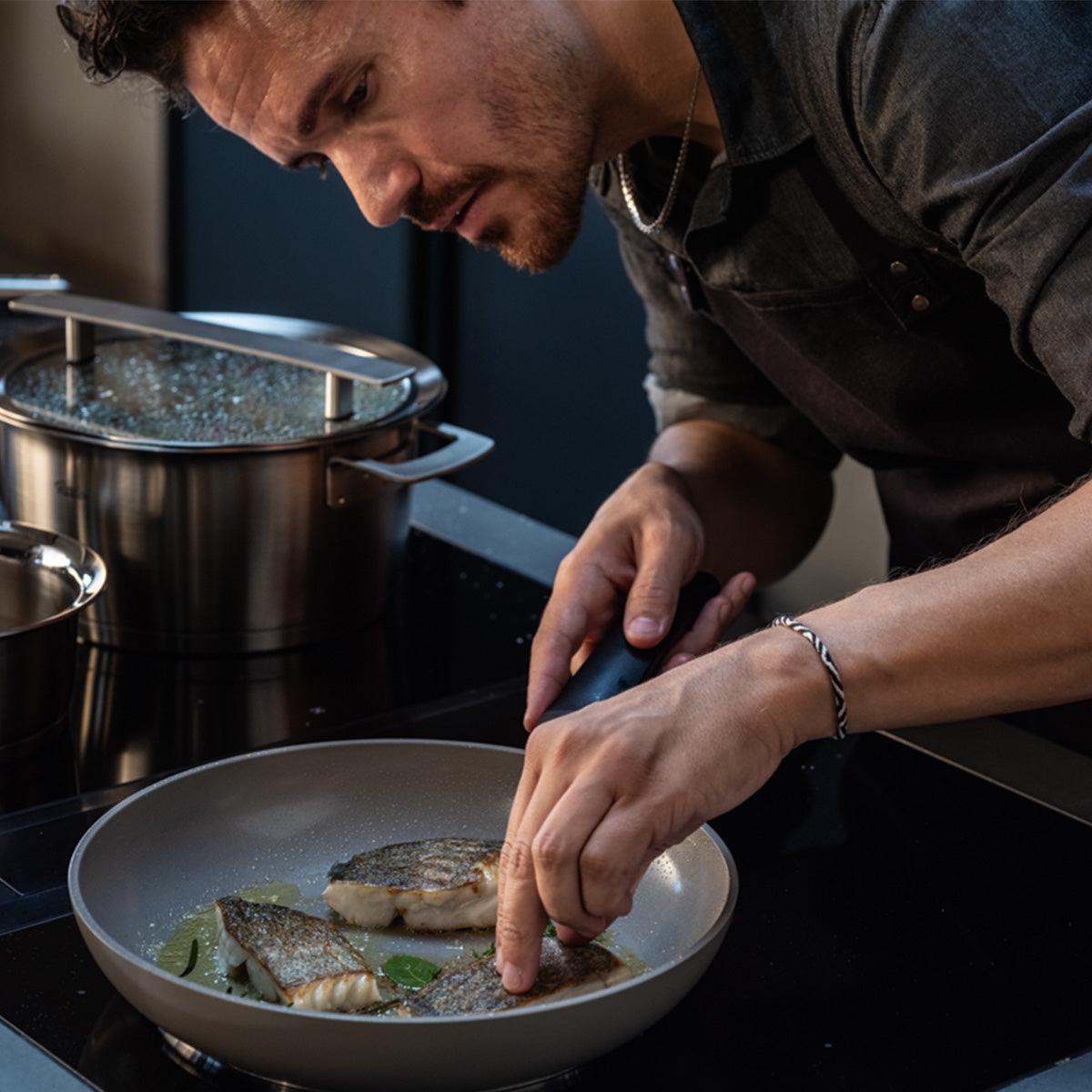 Ein Mann in einem dunklen Hemd und einer Schürze bereitet in einer Fissler Ceratal® Evo Classic Pfanne auf dem Herd gekonnt Fischfilets zu und verfeinert sie zum Schluss mit frischen Kräutern. Die innovative keramische Antihaftbeschichtung der Pfanne sorgt für makellose Ergebnisse, während hochwertige Fissler-Edelstahltöpfe – ein Aushängeschild deutscher Handwerkskunst und deutschen Designs – daneben bereitstehen.
