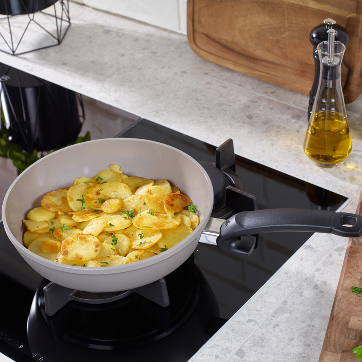 A premium Fissler frying pan with precisely cooked, golden sliced potatoes rests on a stovetop, showcasing the brand’s commitment to German engineering and timeless design. Close by, a glass bottle of olive oil, a pepper grinder, and a wooden cutting board complement the elegant functionality of the kitchen workspace on a light countertop.
