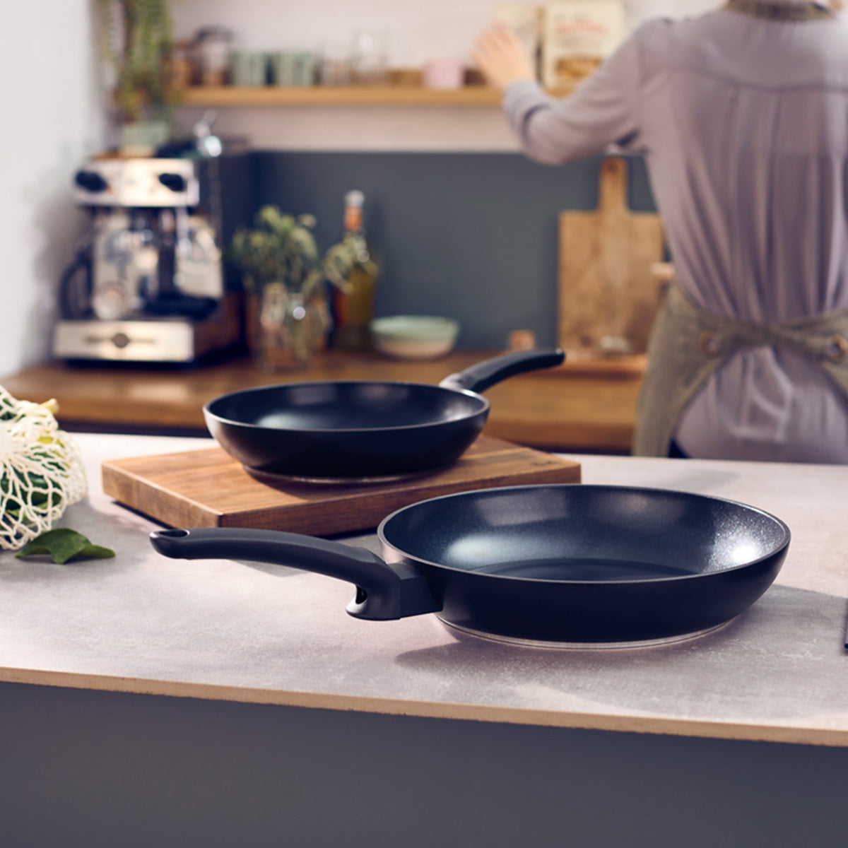 Zwei hochwertige schwarze Fissler Ceratal® Evo Classic-Pfannen mit innovativer Antihaftbeschichtung stehen auf einer Küchenarbeitsplatte, eine davon elegant auf einem Holzschneidebrett platziert. Im sanft unscharfen Hintergrund steht eine Person neben Regalen, die mit hochwertigem Küchenzubehör gefüllt sind – ein Spiegelbild des Engagements von Fissler für Präzisionstechnik und zeitlose deutsche Handwerkskunst seit 1845.