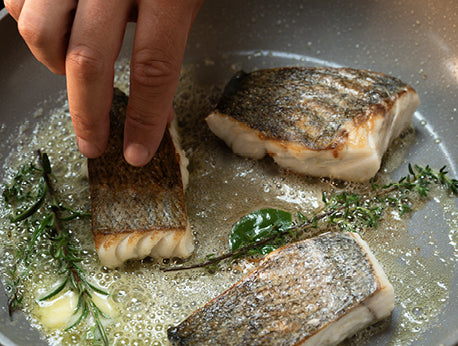 Eine Hand gibt Fisch mit knuspriger Haut in eine Fissler-Pfanne mit sprudelnder Butter und Kräutern und verbindet zwei gleichmäßig garende Filets.