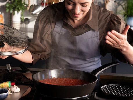 Ein Koch in einer grauen Schürze hebt den Deckel einer Fissler-Pfanne mit roter Soße an, die auf dem Herd inmitten von Küchenkräutern und -utensilien dampft.