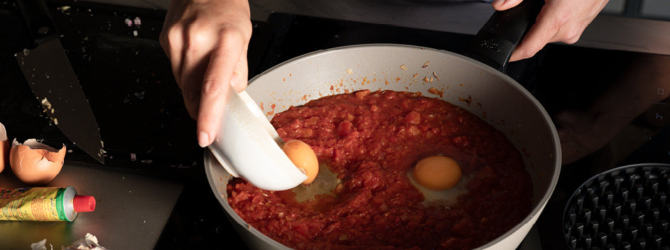 Ein Koch schlägt in einer Fissler-Edelstahlpfanne Eier in Tomatensauce auf; daneben liegen Schalen, Tomatenmark und gehackte Zwiebeln.