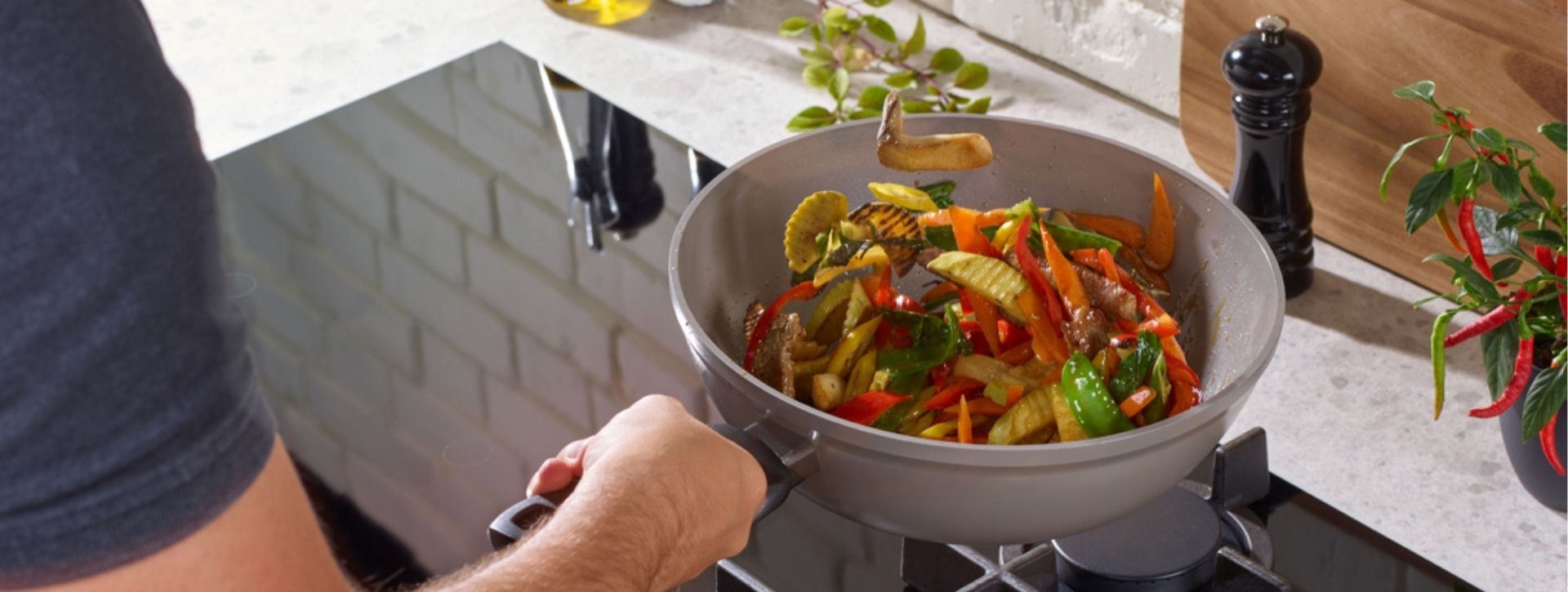 Eine Person bereitet in einer Fissler-Edelstahlpfanne ein lebhaftes Pfannengericht zu, Küchenutensilien und frische Zutaten liegen daneben.
