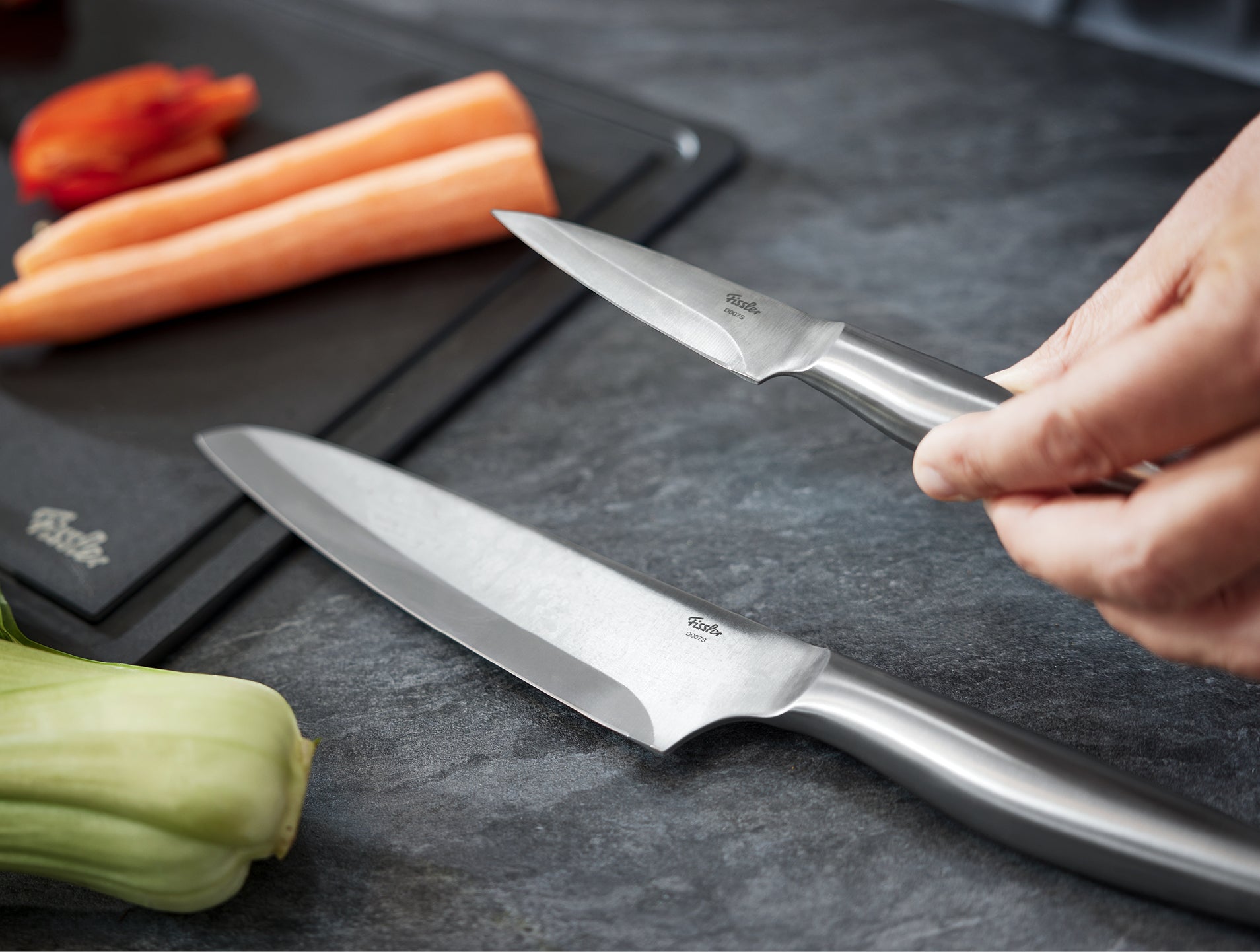 An einer dunklen Theke, auf der Karotten und frisches Gemüse fein säuberlich auf einem Schneidebrett angeordnet sind, arbeitet eine Person mit präzisionsgefertigten Fissler-Messern.