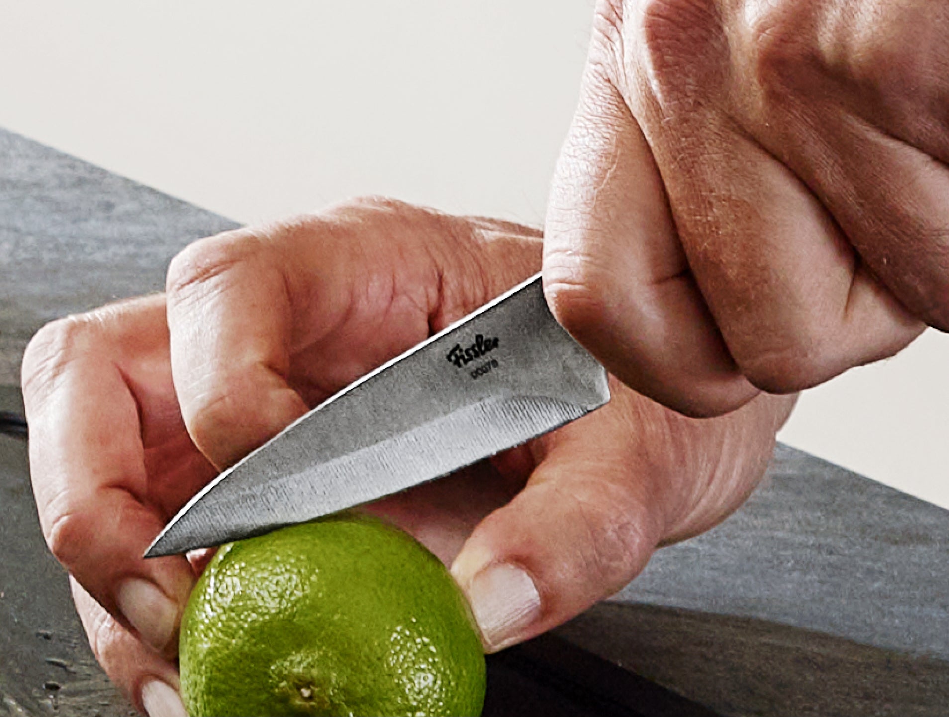 Nahaufnahme einer Hand, die eine grüne Limette hält, während eine andere ein Fissler-Messer aus rostfreiem Stahl benutzt, um sie auf einer dunklen Fläche zu schneiden.