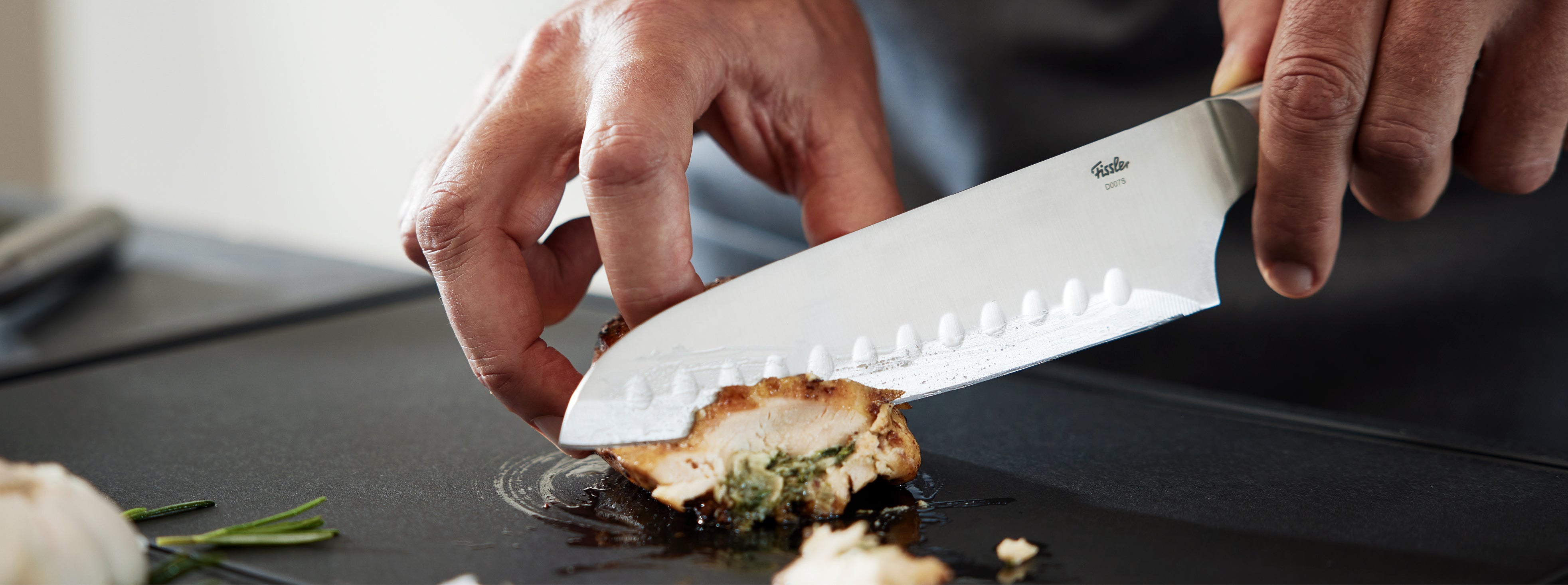 Nahaufnahme eines Kochs, der auf einem Fissler-Schneidebrett mit frischen Kräutern und Knoblauch fachmännisch Hühnerfleisch schneidet und dabei Präzision und Qualität demonstriert.