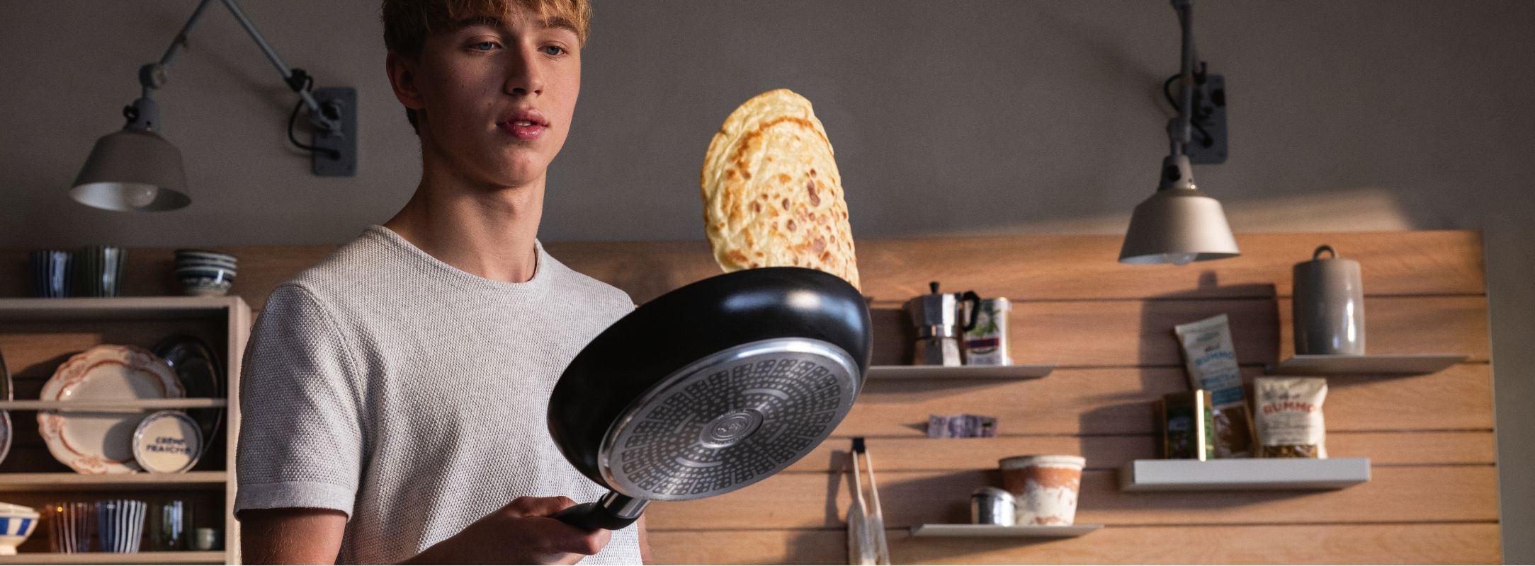 Eine junge Person wendet einen Pfannkuchen in einer Fissler-Pfanne, umgeben von zeitlosem Küchengeschirr und Holzregalen in einer gemütlichen Küche.