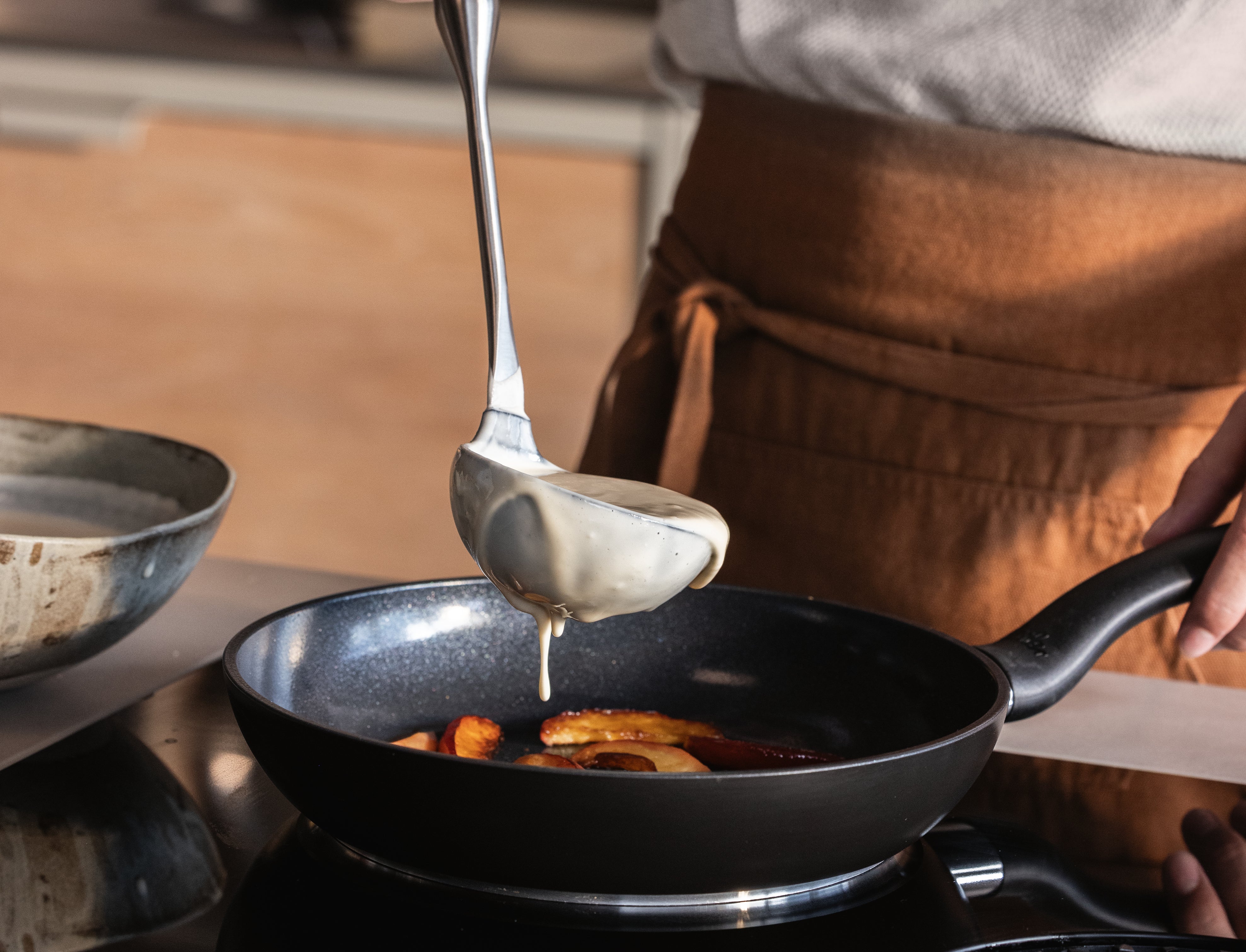 Ein Koch gießt Teig in eine Fissler-Edelstahlpfanne mit Äpfeln auf dem Herd und stellt damit deutsche Innovation und zeitloses Design unter Beweis.