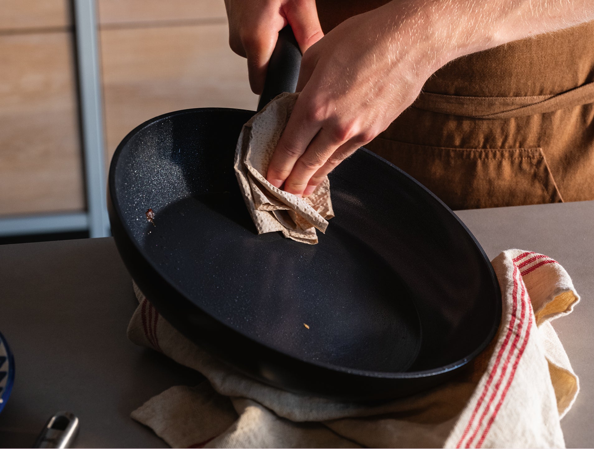 Eine Person wischt sorgfältig eine Fissler-Pfanne auf einer Arbeitsplatte ab, unter der ein rot gestreiftes Handtuch liegt; nur Hände und Oberkörper sind zu sehen.