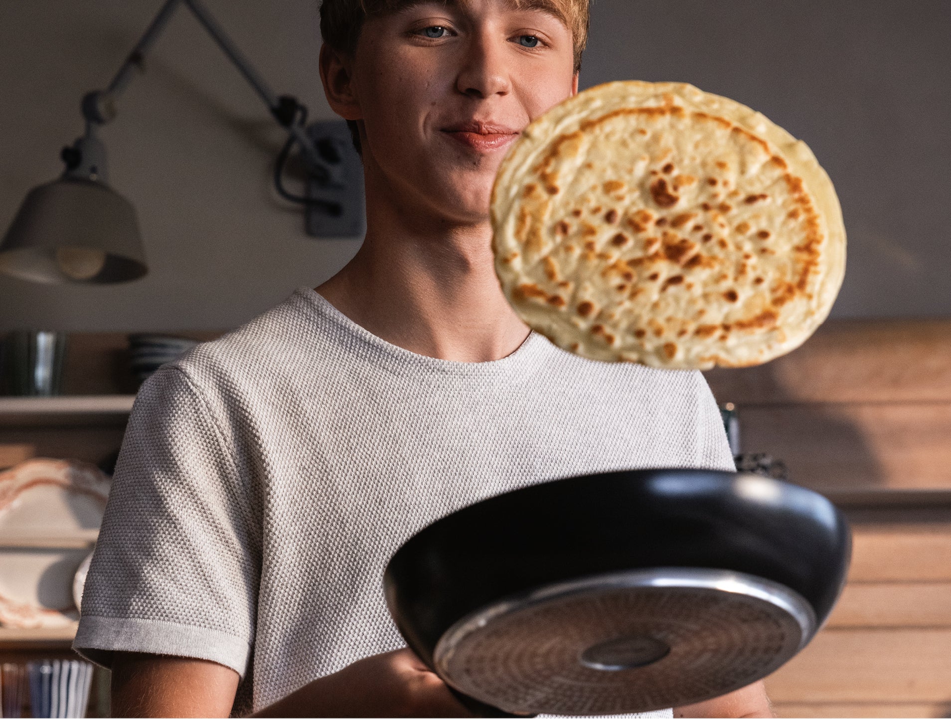 Eine junge Köchin wendet einen Pfannkuchen in der Luft mit einer schwarzen Fissler-Pfanne in einer modernen Küche, hinter der Regale und eine Lampe sanft leuchten.