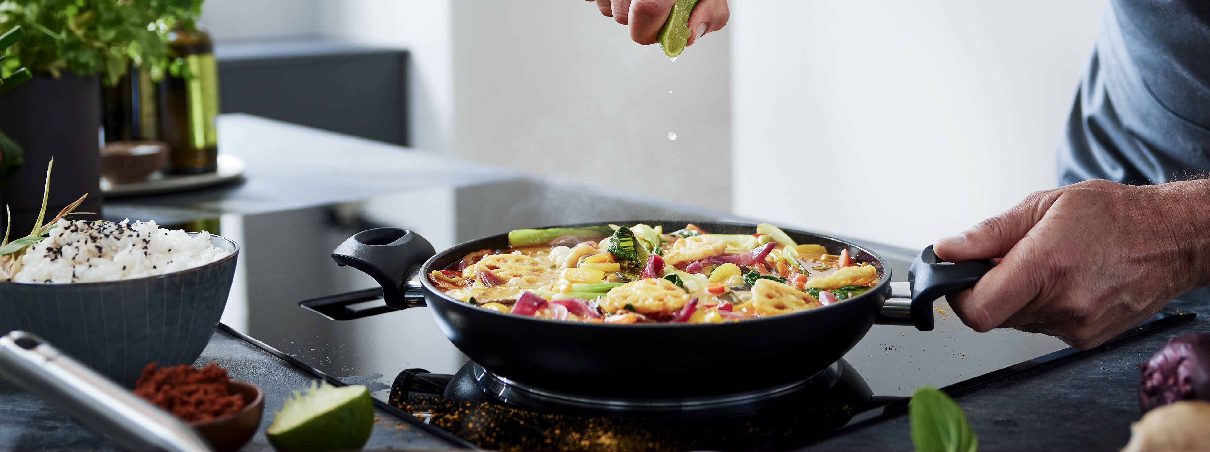 In einer Fissler-Edelstahlpfanne auf einem modernen Herd presst eine Person Limetten über das Gemüse, während Reis und Kräuter in der Nähe sind.
