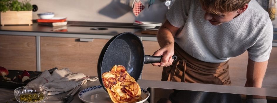 Eine Person benutzt eine Fissler-Pfanne zum Wenden eines Obstpfannkuchens in einer modernen Küche, in der Fissler-Geräte und -Zubehör vorhanden sind.