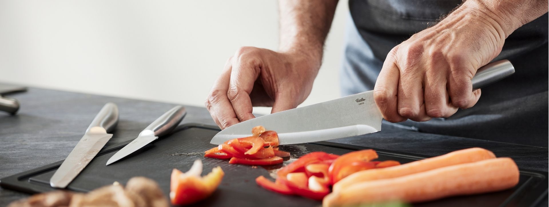 Die Hände einer Person schneiden eine rote Paprika mit einem Fissler-Messer auf einem schwarzen Brett; daneben liegen Karotten, Pilze und weitere Fissler-Messer.
