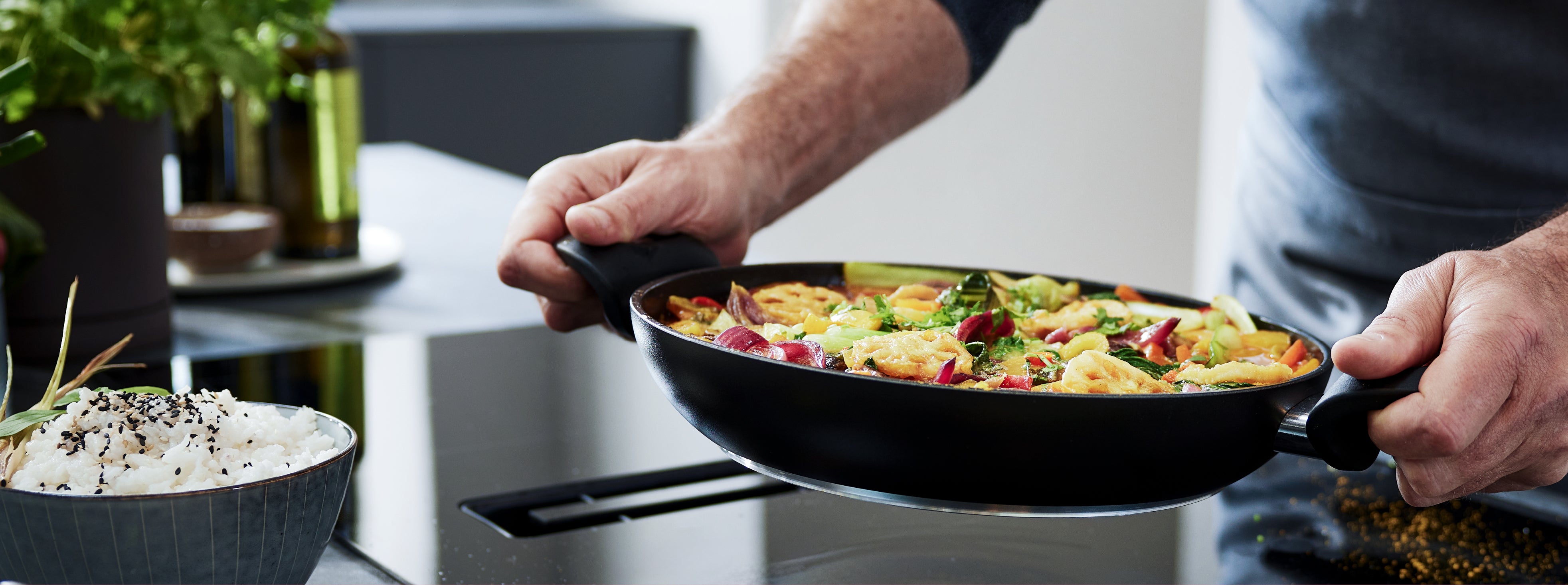 Eine Hand hält eine Fissler-Pfanne mit lebhaft gebratenem Gemüse über einem modernen Herd; daneben liegt Reis mit Sesamkörnern.