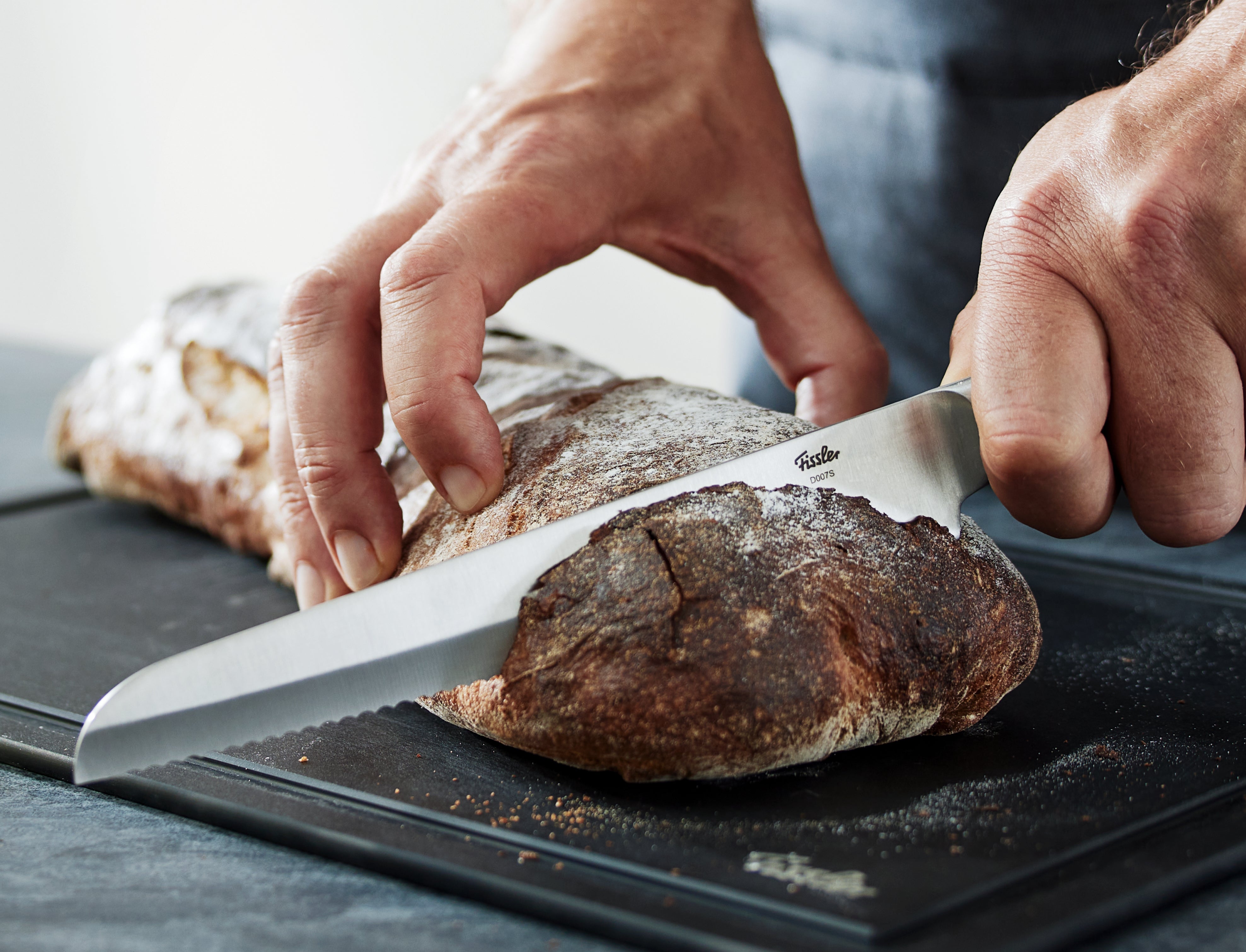 Eine Person schneidet knuspriges Brot auf einem eleganten schwarzen Fissler-Schneidebrett mit einem Präzisions-Zackenmesser, das deutsche Ingenieurskunst demonstriert.