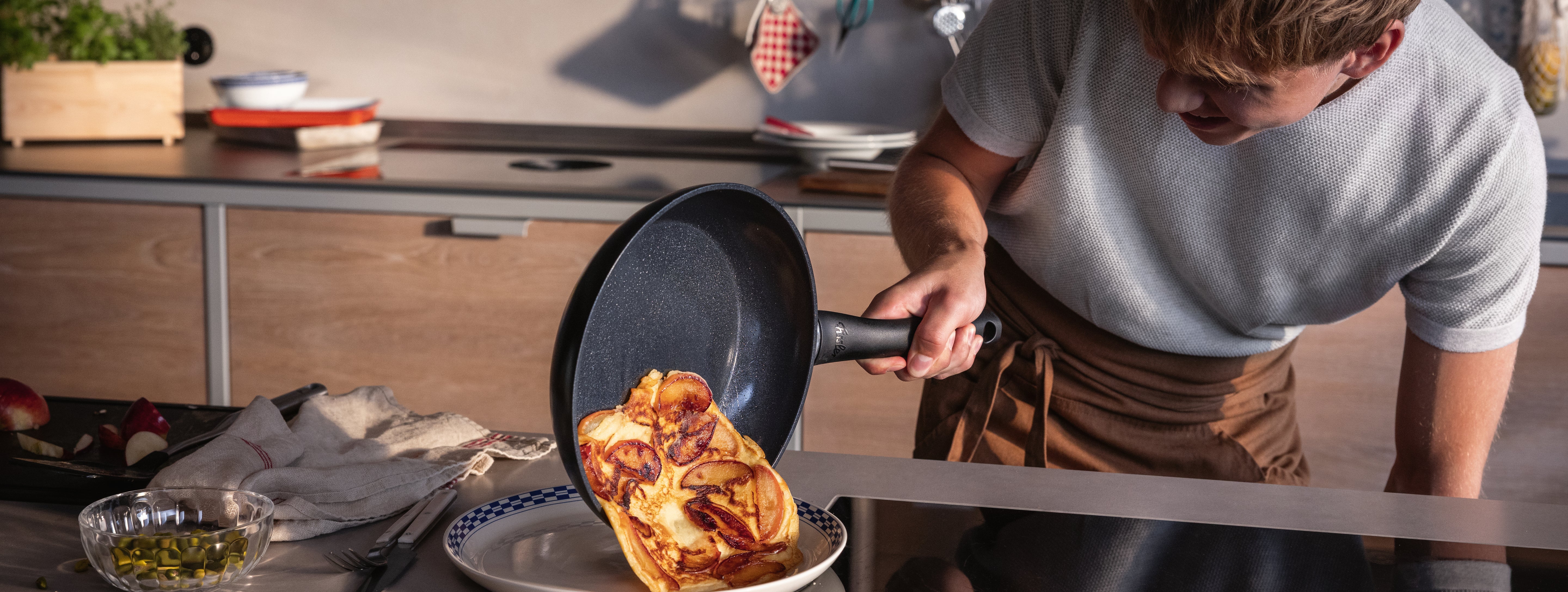 Eine Person schiebt ein goldenes Omelett aus einer Fissler-Pfanne auf einen Teller; hochwertiges Kochgeschirr und frische Zutaten stehen auf der Theke.