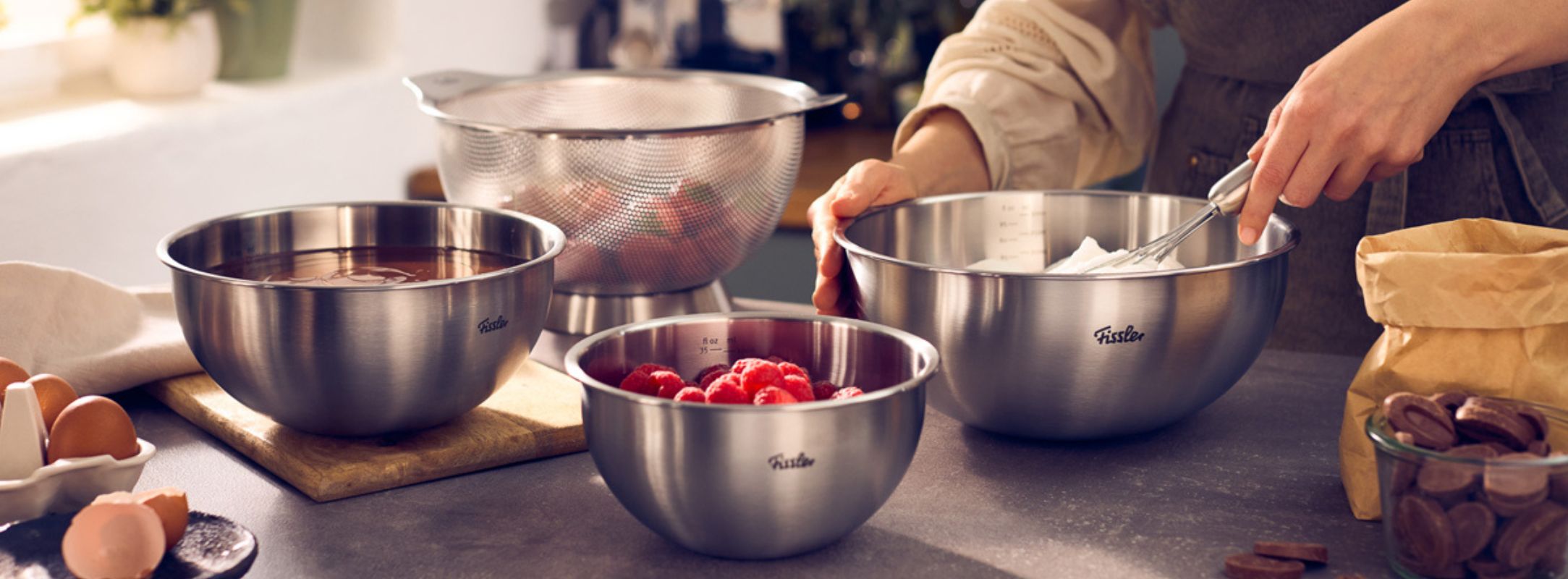 Eine Person bereitet auf einer Küchenarbeitsplatte Zutaten in einer Fissler-Edelstahl-Rührschüssel zu, umgeben von frischen Eiern, reifen Himbeeren, Stückchen feinster Schokolade, einem mit Erdbeeren gefüllten Fissler-Sieb sowie weiteren hochwertigen Fissler-Rührschüsseln aus Metall – ein Beispiel für präzise gefertigte Kochgeschirrprodukte, die auf außergewöhnliche Leistungsfähigkeit und Langlebigkeit ausgelegt sind.