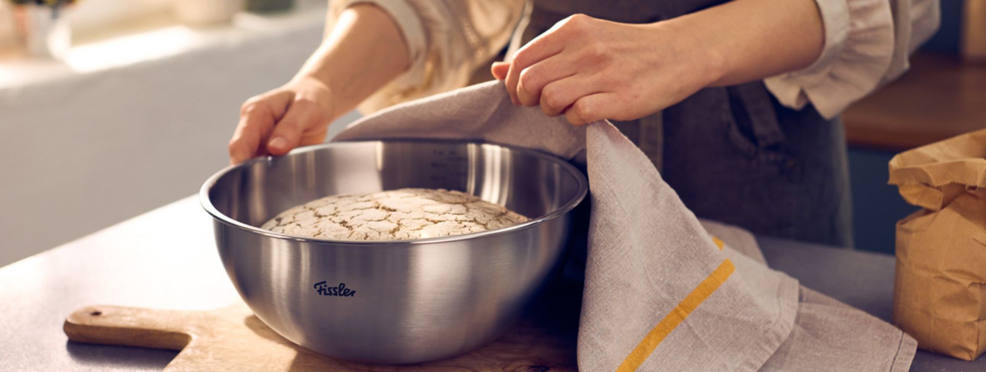 Eine Person hebt ein Tuch an und gibt den Blick auf perfekt aufgegangenen Teig in einer Fissler-Edelstahlschüssel auf einer Küchenarbeitsplatte frei. Daneben liegen Mehl und eine Papiertüte, die die Präzision und Sorgfalt beim Brotbacken zu Hause mit Fisslers traditionellem Qualitätskochgeschirr unterstreichen.
