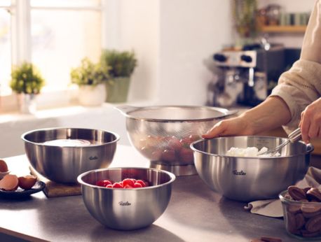 In einer hell erleuchteten Küche verrührt eine Person mühelos Zutaten in einer Fissler-Edelstahlschüssel und verkörpert damit deutsche Handwerkskunst. In drei weiteren Fissler-Metallschüsseln sind Eier, Erdbeeren und Schlagsahne elegant angerichtet. Sonnenlicht strömt durch das Fenster und beleuchtet hochwertiges Kochgeschirr und Küchengeräte, die Fisslers Tradition von Präzisionstechnik und zeitlosem Design verkörpern.