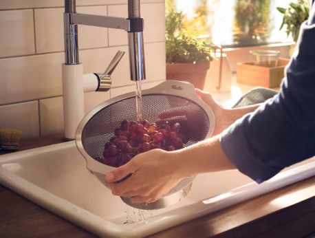 Eine Person spült eine Traube roter Weintrauben in einem hochwertigen Fissler-Edelstahlsieb unter fließendem Wasser am Spülbecken ab, während natürliches Sonnenlicht im Hintergrund das zeitlose Design und die erstklassige Verarbeitung des in Deutschland hergestellten Kochgeschirrs hervorhebt.
