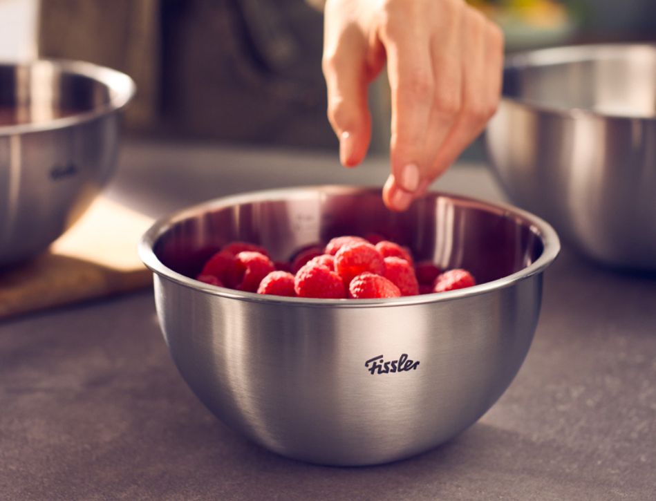 Eine Hand greift in eine hochwertige Fissler-Rührschüssel aus Edelstahl, die mit frischen Himbeeren gefüllt ist und auf einer eleganten grauen Arbeitsplatte steht – ein Beispiel für Fisslers präzise verarbeitete, in Deutschland hergestellte Handwerkskunst und sein zeitloses Design.