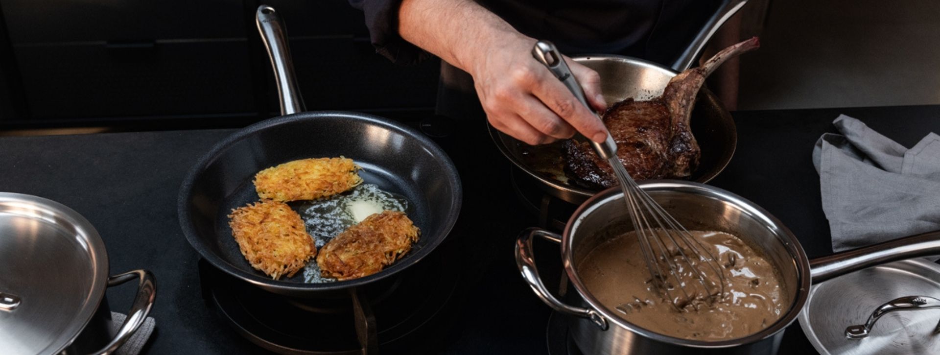 Ein Koch verwendet Fissler Edelstahlpfannen und -töpfe, um Kartoffelkuchen, Soße und Steak zuzubereiten; daneben liegen Utensilien und eine graue Serviette.