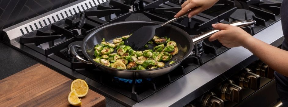 Eine Person sautiert Rosenkohl in einer Fissler-Pfanne auf einem Gasherd; eine halbierte Zitrone liegt auf einem Holzbrett daneben.