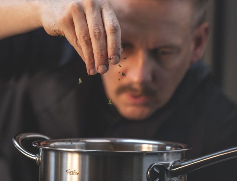 Eine Köchin streut Kräuter in einen Fissler-Edelstahltopf, der deutsche Ingenieurskunst und zeitloses Design in der Küche demonstriert.