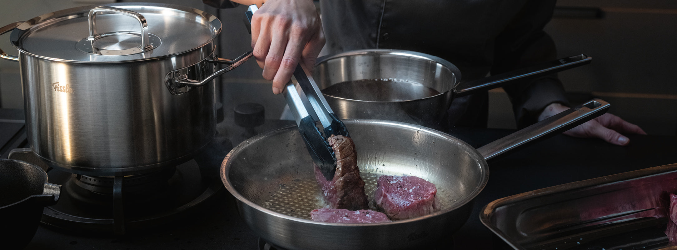 Eine Person brät Fleisch mit einer Zange in einer Fissler-Edelstahlbratpfanne an, die neben unserem charakteristischen Topf und der Kasserolle auf dem Herd steht.