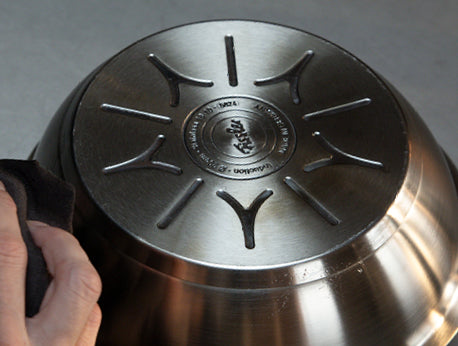 Eine Hand wischt über den Boden einer Fissler-Edelstahlschüssel und enthüllt dabei das präzise eingravierte Logo und die Markierungen, die auf dem Kopf stehen und grau sind.