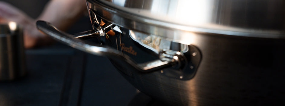 Nahaufnahme einer Fissler-Edelstahlkanne mit präzisionsgefertigtem Griff, die Licht reflektiert. Unscharfe Hand und Tasse im Hintergrund.