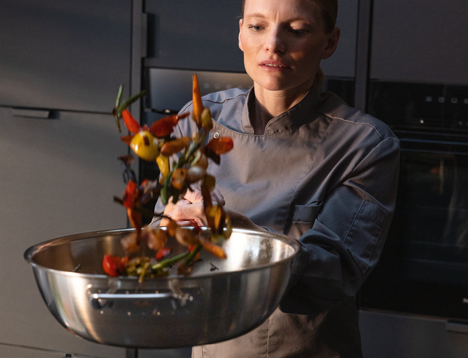 Ein Chefkoch in Grau schwenkt mit einer Fissler-Edelstahlpfanne lebhaftes Gemüse und stellt damit Präzision und Qualität in einer modernen Küche unter Beweis.