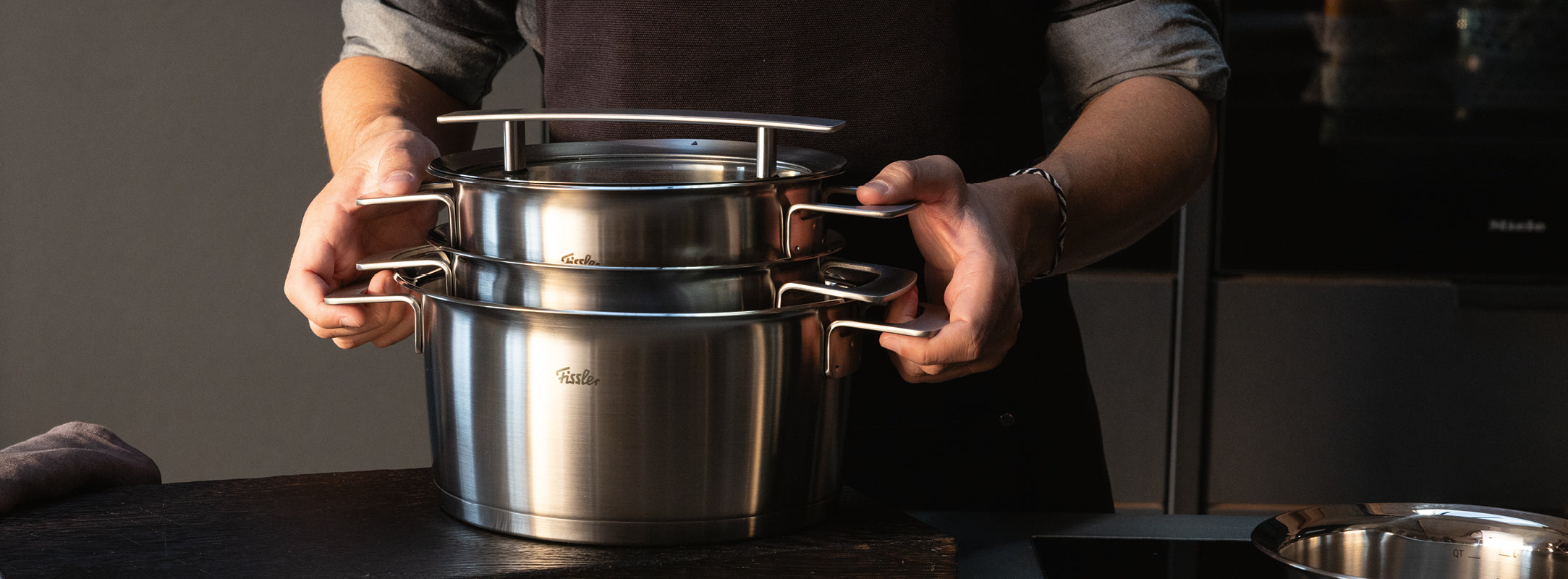 Eine Person stapelt die in Deutschland hergestellten Fissler-Edelstahltöpfe mit ihren haltbaren Griffen und der zeitlosen, polierten Oberfläche auf einer Küchentheke.