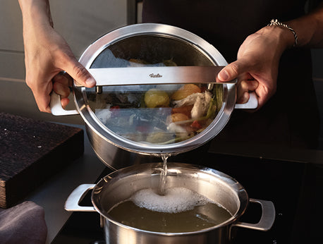 Ein Hausmann gießt Brühe aus einem Fissler-Edelstahltopf und benutzt den Präzisionsdeckel, um die festen Bestandteile in einen anderen Topf abzusieben.