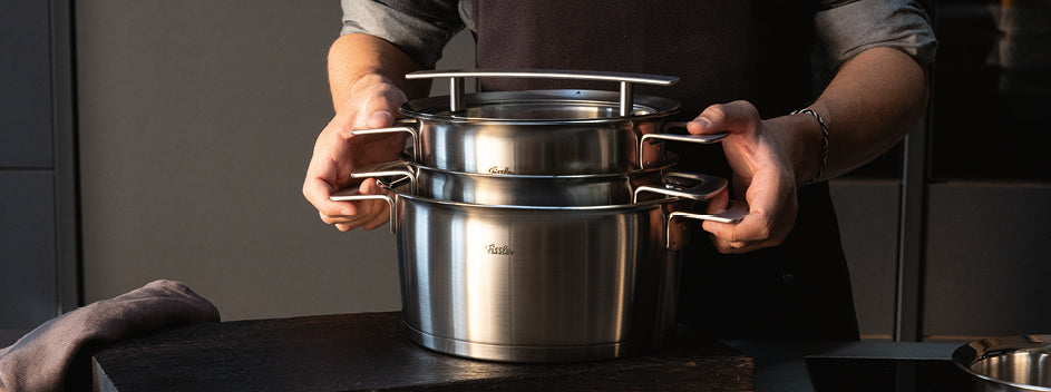 Eine Person stapelt zwei Fissler-Edelstahltöpfe mit Deckeln auf einer dunklen Arbeitsplatte in einer modernen Küche, die deutsches Präzisionsdesign zeigt.