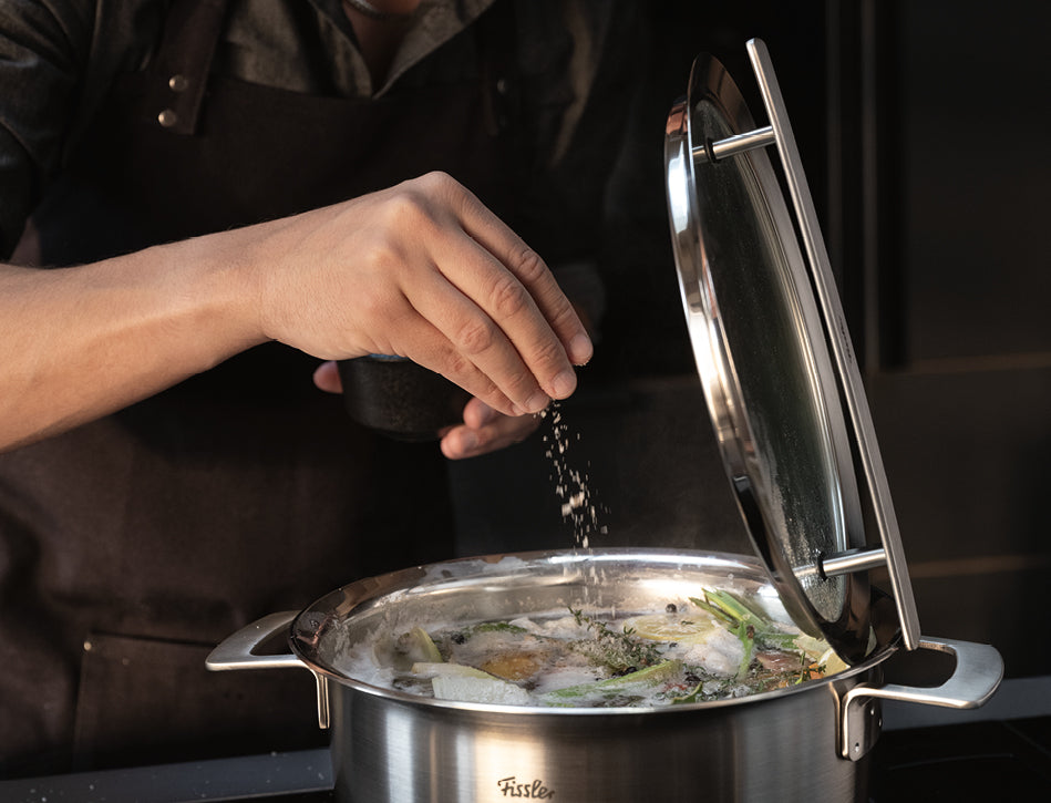 Ein Koch würzt ein köchelndes Gericht in einem Fissler-Edelstahltopf und hebt den Deckel an, um die frischen Zutaten in einer modernen Küche freizulegen.
