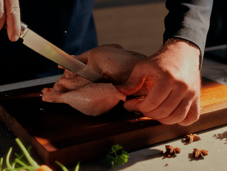 Eine Person tranchiert fachmännisch ein rohes ganzes Huhn mit einem Fissler-Kochmesser auf einem Holzbrett, frische Kräuter und Gemüse in der Nähe.
