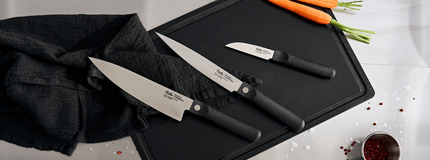 Fissler Präzisionsküchenmesser auf einem schwarzen Brett, daneben Karotten, Handtuch, Salz, Pfefferkörner und eine kleine Tasse mit rotem Gewürz.
