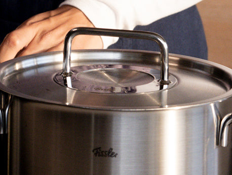 Eine Hand ruht auf dem Deckel eines Fissler-Edelstahltopfes und unterstreicht die hochwertige deutsche Handwerkskunst und den robusten, eleganten Griff.