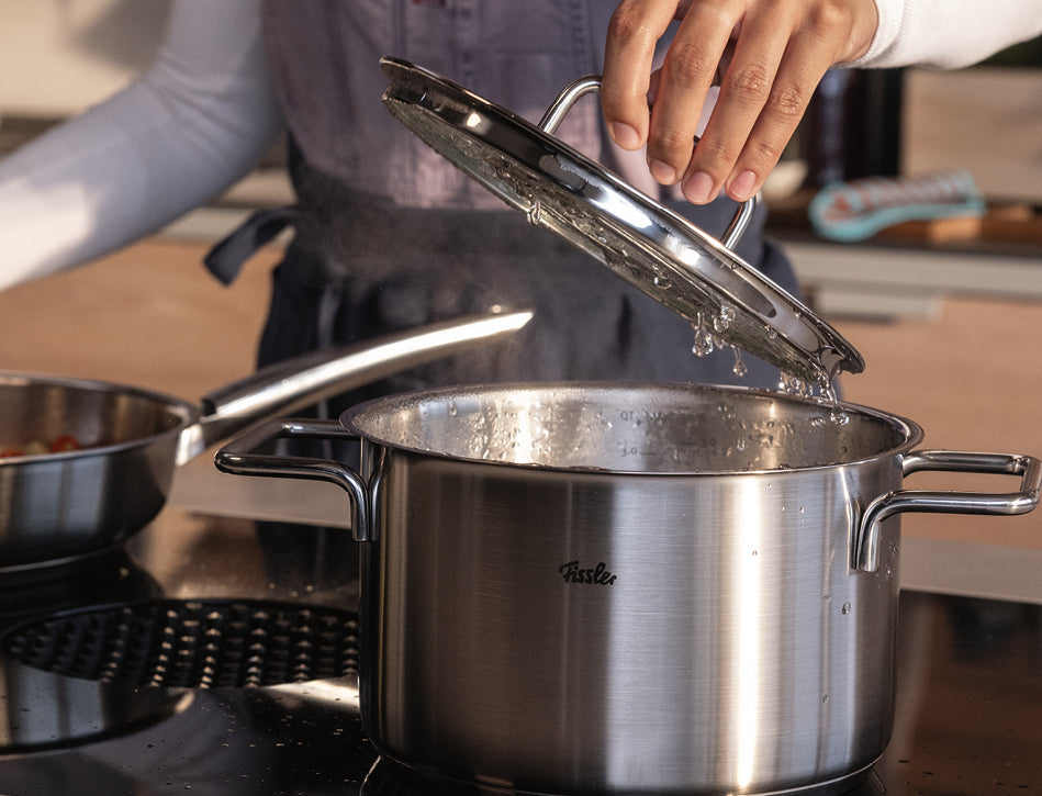 Eine Person hebt den Deckel von einem dampfenden Fissler-Edelstahltopf ab, der durch sein präzises Design besticht, und setzt einen weiteren hochwertigen Topf dahinter.