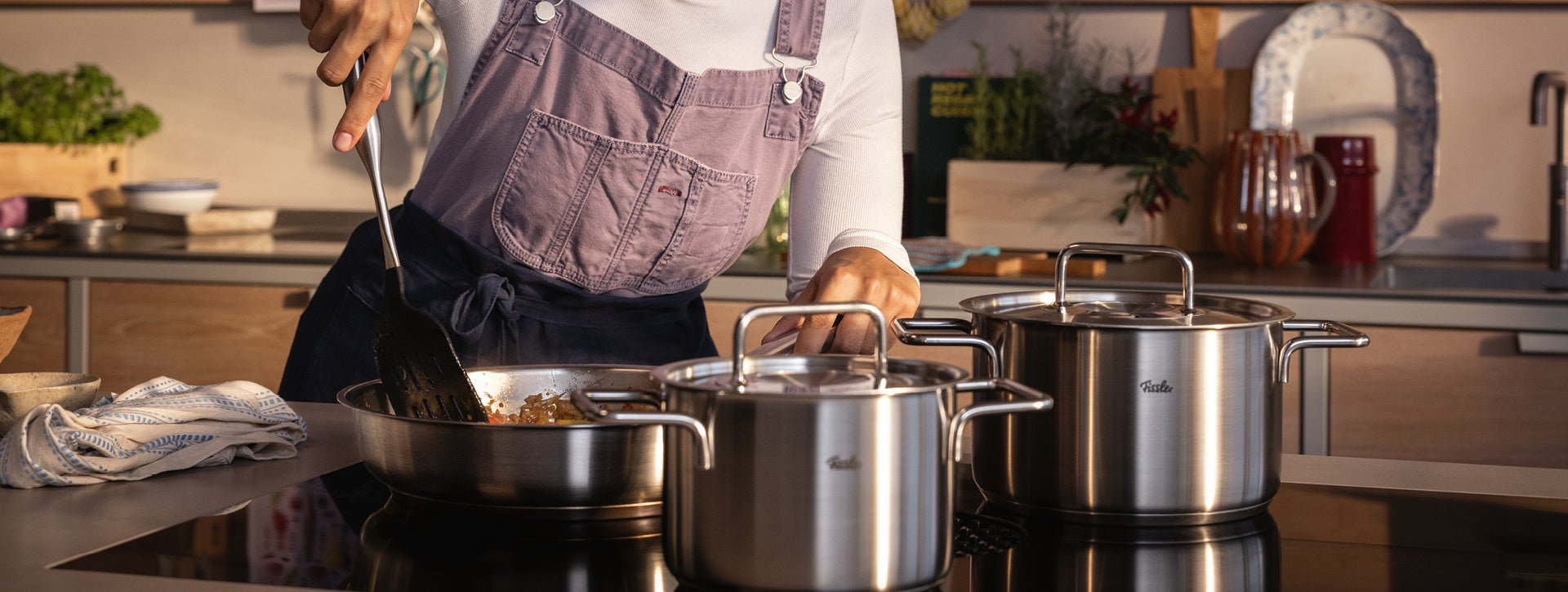 Eine Köchin in rosa Schürze kocht mit Fissler-Edelstahltöpfen auf dem Herd; Grünzeug und hochwertiges Kochgeschirr schmücken die Küche.