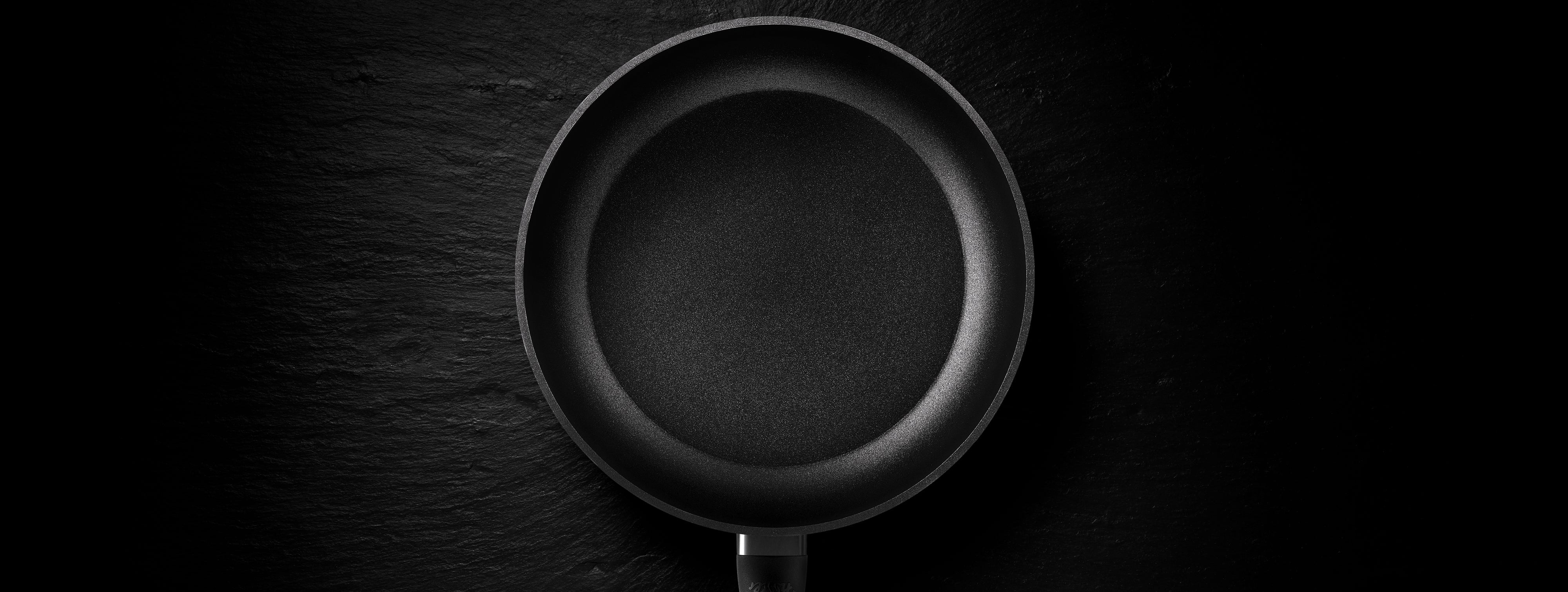 Eine schwarze antihaftbeschichtete Bratpfanne von Fissler, von oben betrachtet auf einem dunkel strukturierten Hintergrund, zeigt ihre präzisionsgefertigte Oberfläche.