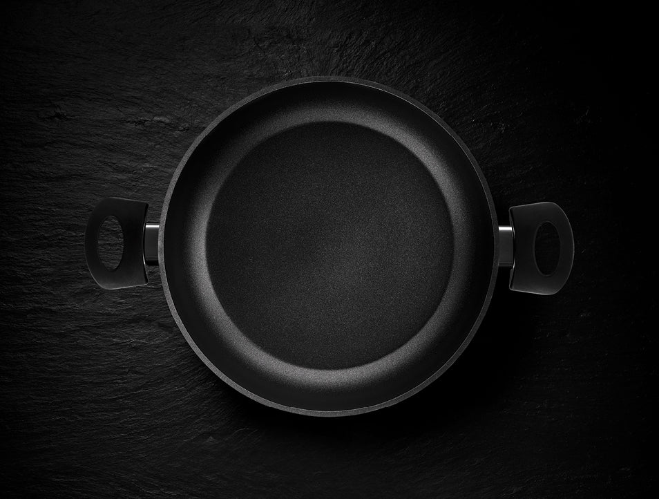 Eine schwarze, antihaftbeschichtete Pfanne von Fissler mit zwei ergonomischen Griffen, von oben gesehen auf einer dunklen Oberfläche; Präzisionsarbeit in Deutschland.